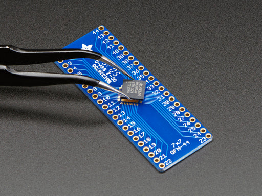 A pair of tweezers holding a microchip hovers over the SMT Breakout PCB for 44-QFN or 44-TQFP placement on a long blue rectangular breakout board.