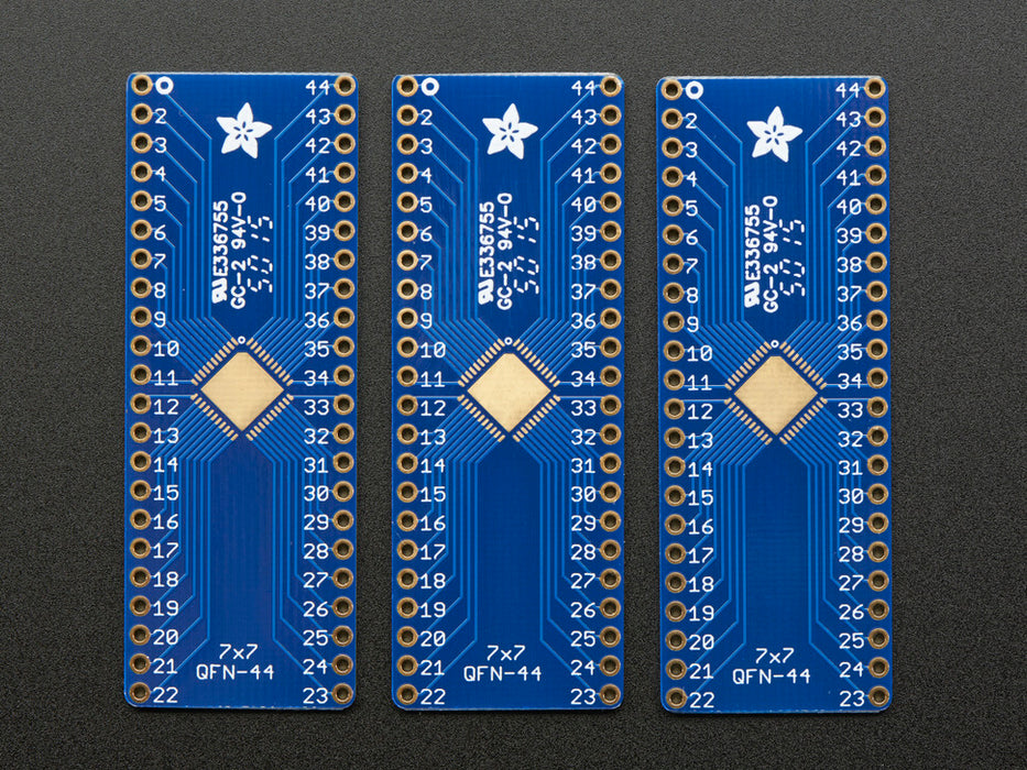 A pair of tweezers holding a microchip hovers over the SMT Breakout PCB for 44-QFN or 44-TQFP placement on a long blue rectangular breakout board.