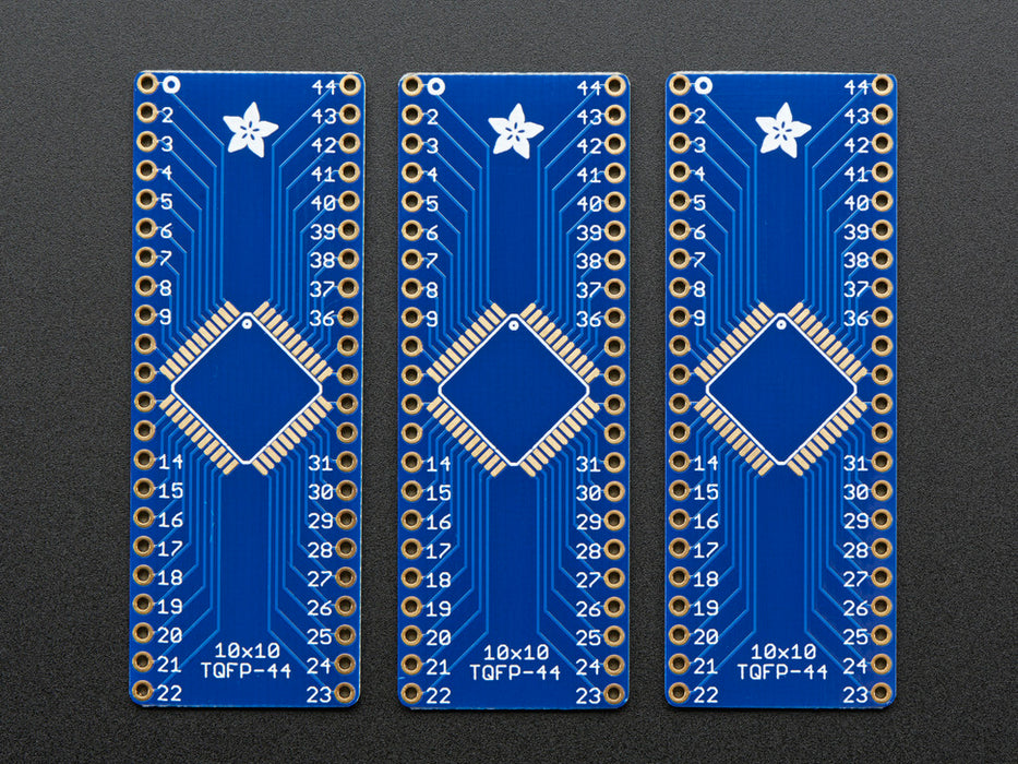 A pair of tweezers holding a microchip hovers over the SMT Breakout PCB for 44-QFN or 44-TQFP placement on a long blue rectangular breakout board.