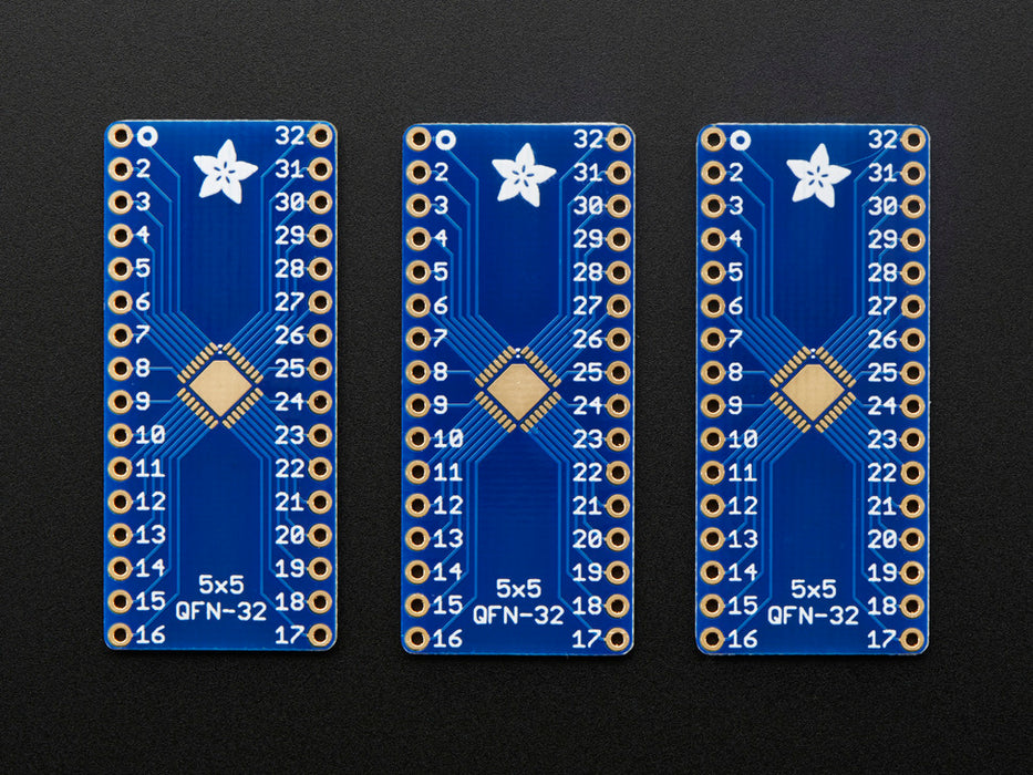 A pair of tweezers holding a microchip hovers over the SMT Breakout PCB for 32-QFN or 32-TQFP placement on a long blue rectangular breakout board.