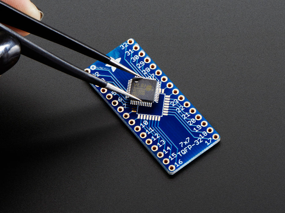 A pair of tweezers holding a microchip hovers over the SMT Breakout PCB for 32-QFN or 32-TQFP placement on a long blue rectangular breakout board.