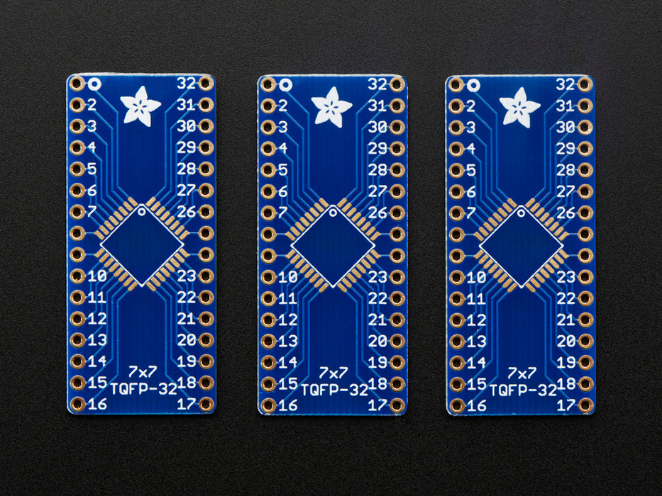 A pair of tweezers holding a microchip hovers over the SMT Breakout PCB for 32-QFN or 32-TQFP placement on a long blue rectangular breakout board.