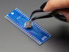 A pair of tweezers holding a microchip hovers over the SMT Breakout PCB for 48-QFN or 48-TQFP placement on a long blue rectangular breakout board.