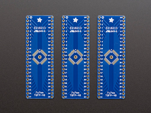 A pair of tweezers holding a microchip hovers over the SMT Breakout PCB for 48-QFN or 48-TQFP placement on a long blue rectangular breakout board.