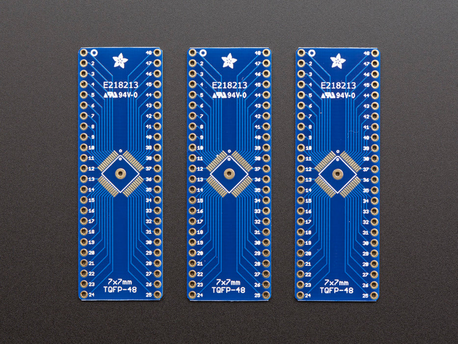 A pair of tweezers holding a microchip hovers over the SMT Breakout PCB for 48-QFN or 48-TQFP placement on a long blue rectangular breakout board.