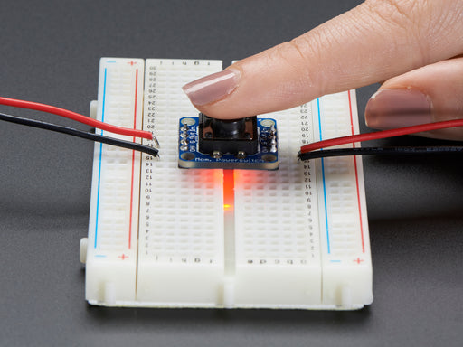 Adafruit Push-button Power Switch Breakout Board plugged into a full-size breadboard and a white person's finger pressing on the button.