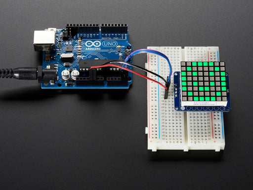 Close-up of soldered and assembled Small 1.2" 8x8 Ultra Bright Square Green LED Matrix + Backpack on a breadboard powered by an Arduino Uno. The LED Matrix displays a green smiley.