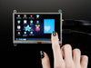 Black polished finger touching the display of a HDMI 5" Display Backpack. The HDMI screen displays a desktop image including the Adafruit logo, the Raspberry Pi logo, and a pink heart.