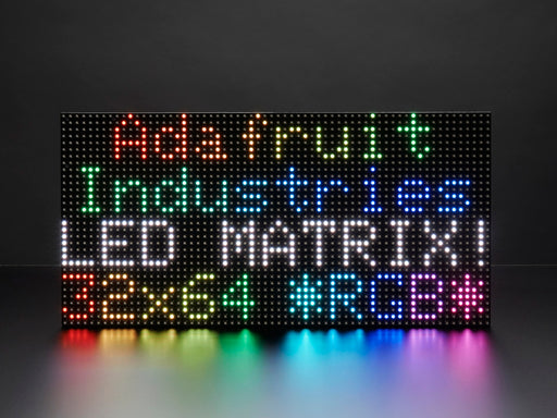 Two white hands hold out an assembled and powered on 64x32 RGB LED Matrix Panel - 6mm pitch. The matrix displays "Adafruit Industries LED MATRIX! 32x64 *RGB*"