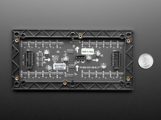 Two white hands hold out an assembled and powered on 64x32 RGB LED Matrix Panel - 6mm pitch. The matrix displays "Adafruit Industries LED MATRIX! 32x64 *RGB*"