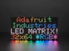 Two white hands hold out an assembled and powered on 64x32 RGB LED Matrix Panel - 5mm pitch. The matrix displays "Adafruit Industries LED MATRIX! 32x64 *RGB*"