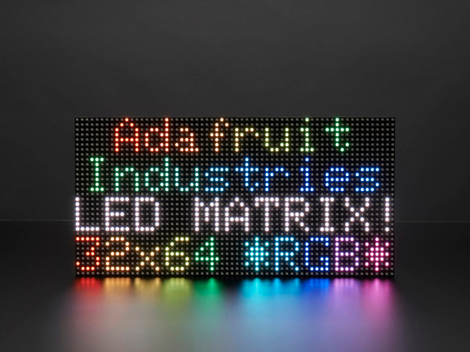 Two white hands hold out an assembled and powered on 64x32 RGB LED Matrix Panel - 5mm pitch. The matrix displays "Adafruit Industries LED MATRIX! 32x64 *RGB*"