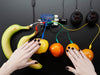 Video of a white hand pressing various bananas, oranges, and other fruits connected to a Raspberry Pi with Adafruit Capacitive Touch HAT via alligator test clip leads.