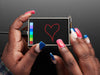 Overhead shot of a Black woman's hands with a blue and pinkish-red manicure drawing a heart on a touchscreen breakout.