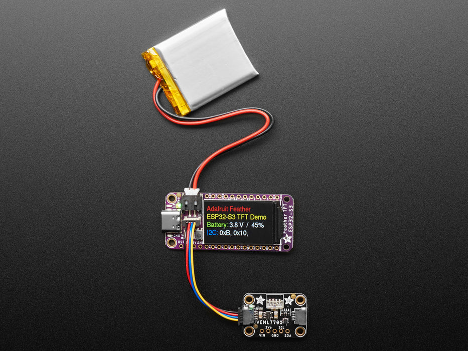 Top view of a purple rectangular microcontroller with a color TFT screen and powered by an ESP32-S3 chip. It is also connected to a Li-Poly battery and a lux sensor breakout. The TFT display reads in colored text: "Adafruit Feather
ESP32-S3 TFT Demo
Battery: 3.8V / 45%
I2C: 0xB, 0x10, "