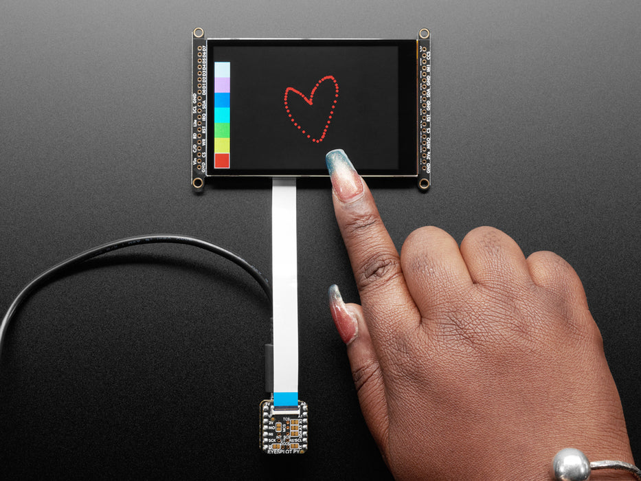 Overhead shot of a Black woman's green-manicured hand touching a 3.5" TFT display breakout connected to a small, square-shaped microcontroller. The TFT screen displays a drawn red heart.